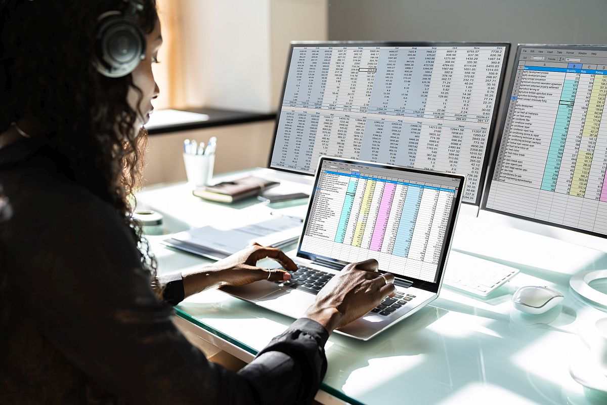 Woman working on laptop with two large monitors, all filled with spreadsheets; synthetic data concept