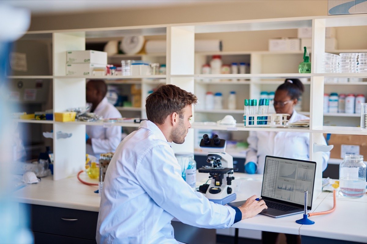 Technician working with a microscope and entering data into a laptop while sitting at a table in a lab; data silos concept