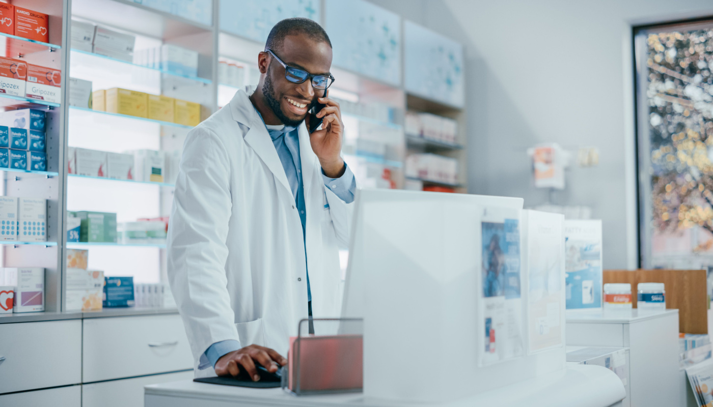 Smiling pharmacist on the phone, using computer behind the pharmacy counter; clinical trial enrollment concept